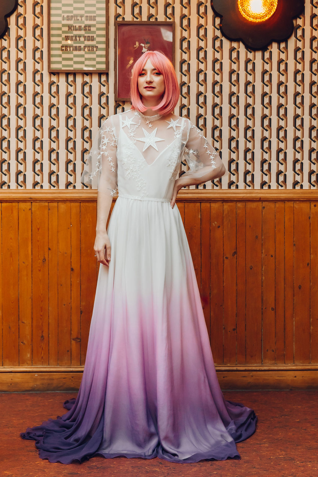 A person wearing a dip-dyed wedding gown with a white top and a gradient from white to pink to purple on the bottom. They are wearing a celestial wedding cape decorated with stars.