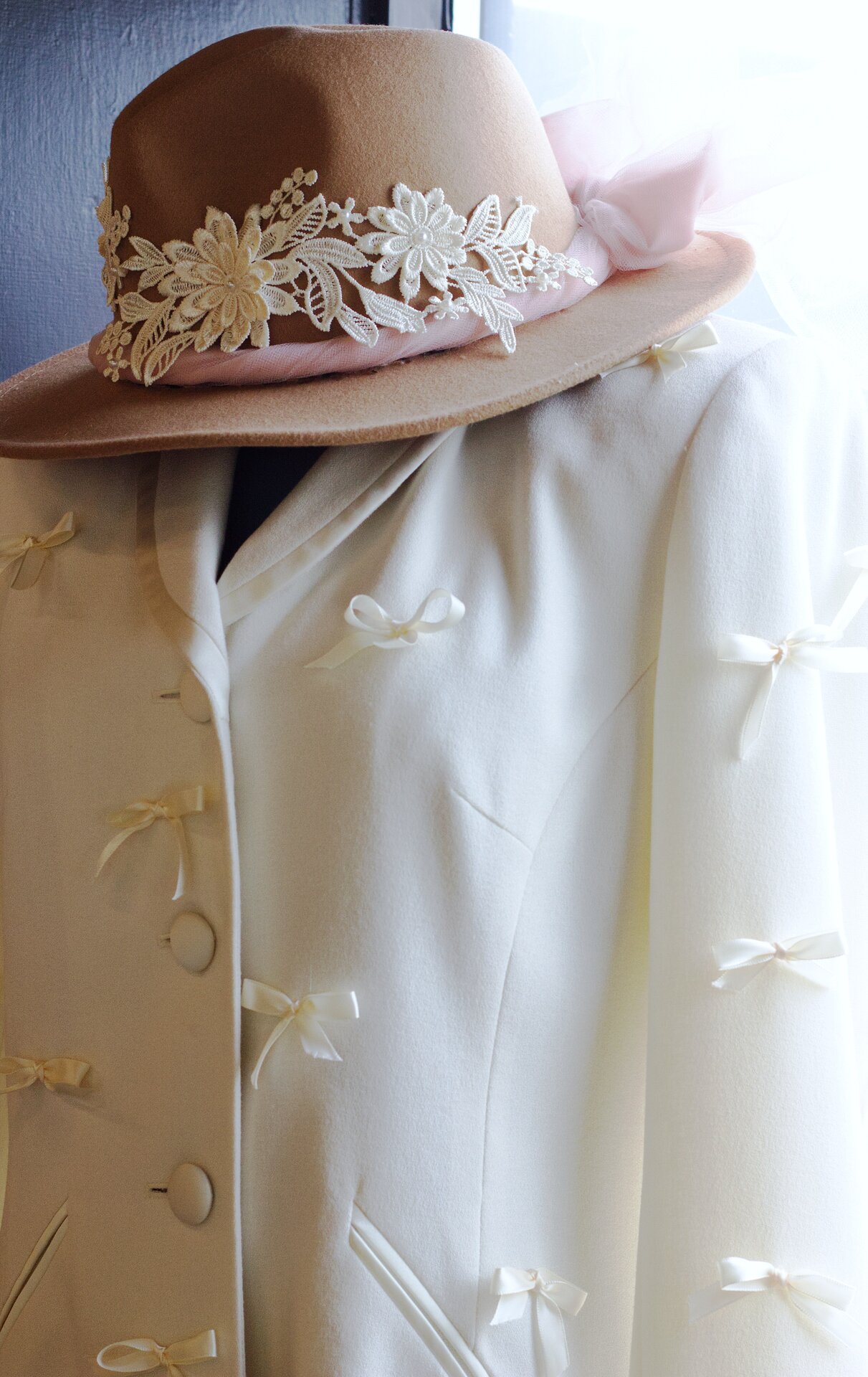 A handmade brown cowboy hat with lace detail and a tulle bow, paired with a handmade white women's blazer decorated with bows.