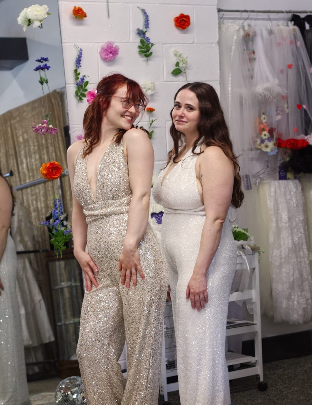 Two brides modeling together wearing sequined bridal jumpsuits in white and champagne.