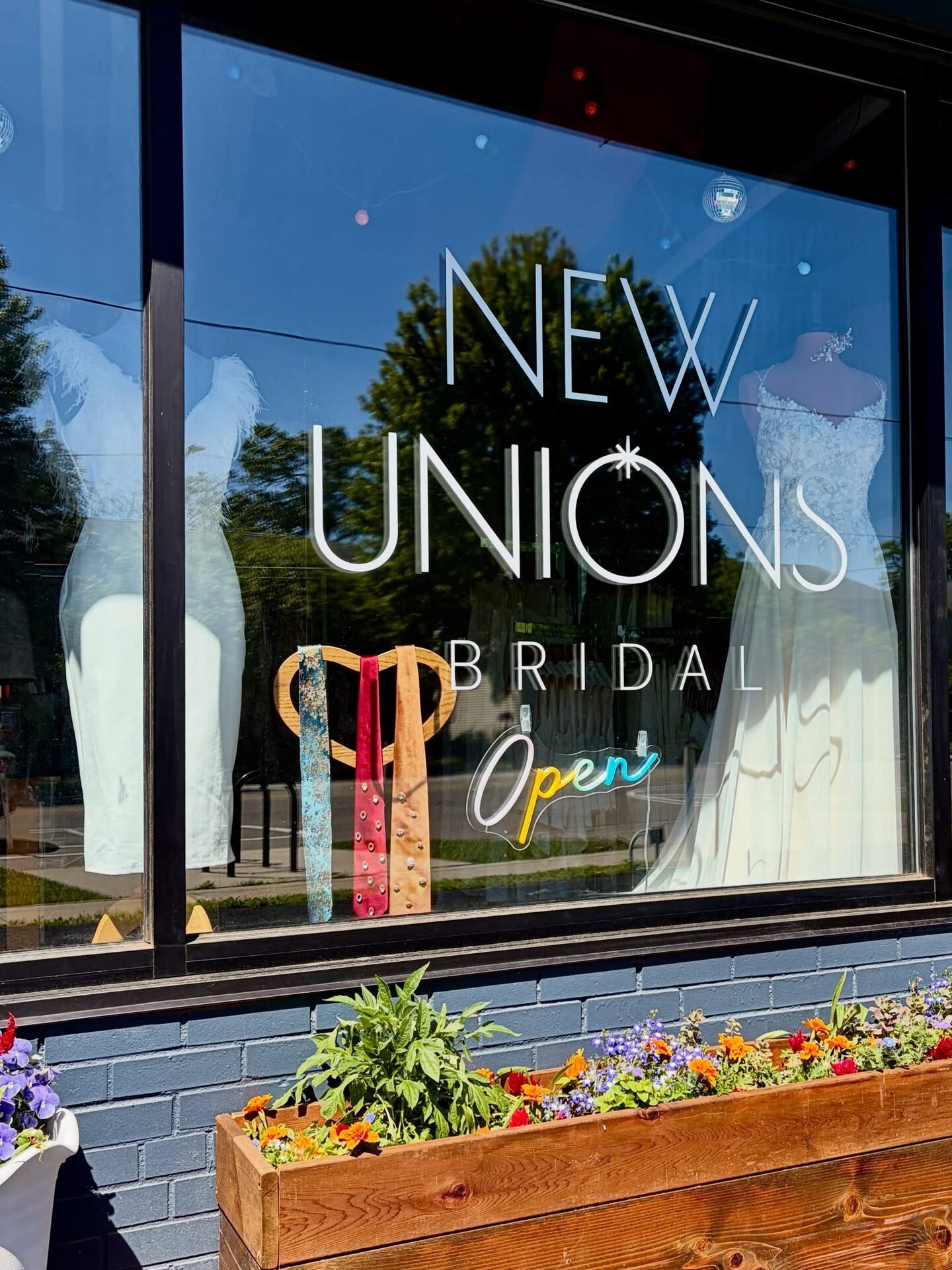 Exterior of the New Unions Bridal storefront. Some wedding dresses and colorful rhinestone neckties can be seen on display in the window.