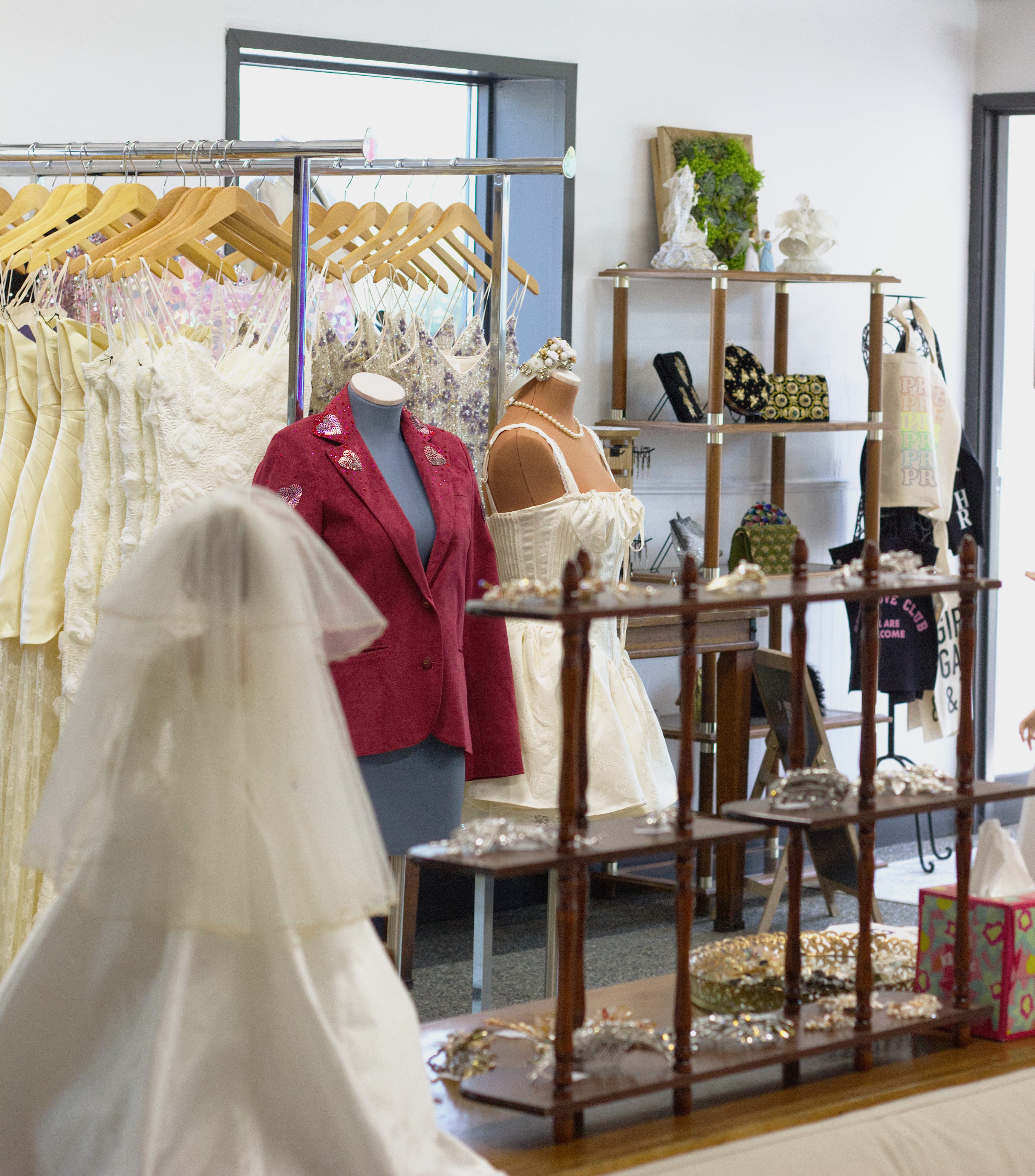 A view of the store interior, showing a white dress and red suit jacket on mannequins, a rack of wedding attire, and some shelves with accessories on them.