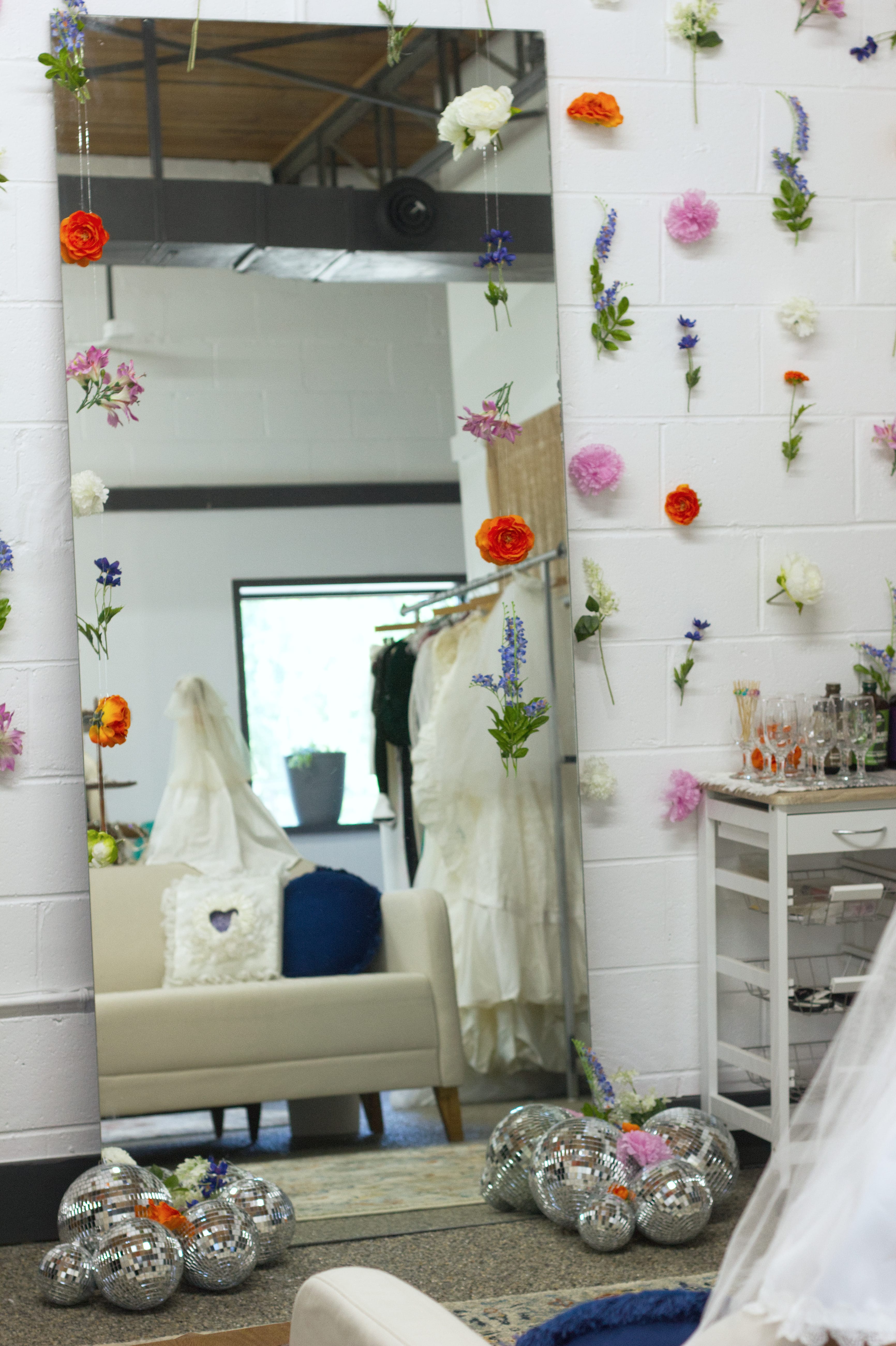 A large mirror in front of a couch, surrounded by disco balls on the floor and strings of flowers hung on the wall around it.