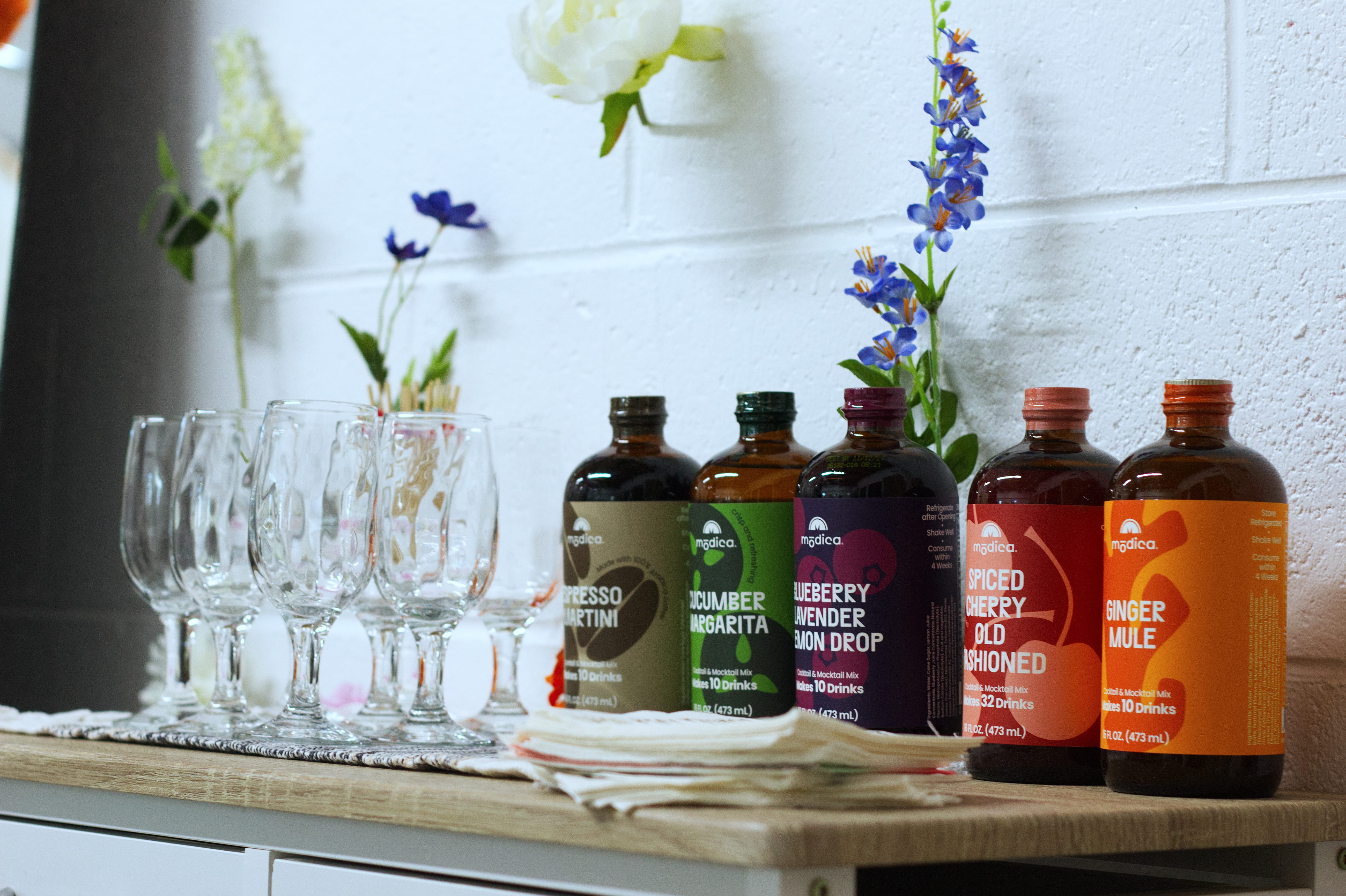 A bar cart with a set of glasses and a variety of colorful bottles of mocktail mixes.